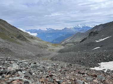 Felsiges Bergtal mit verstreuten Steinen, das zu entfernten schneebedeckten Alpengipfeln unter bewölktem Himmel beim Aufstieg zum Augustbordpass führt.