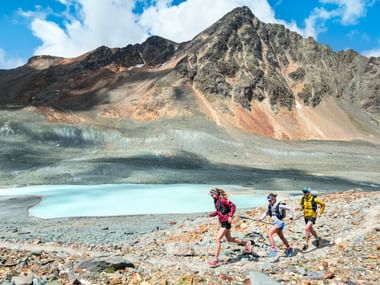 Trois coureurs de trail font du jogging sur un chemin de pierre à côté d'un lac de montagne turquoise sur la Via Grischuna, avec des pics rocheux sous un ciel bleu.