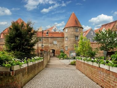 Steinbrücke mit Blumenkästen führt zu mittelalterlichem Rundturm und historischen Gebäuden in Donauwörth unter blauem Himmel mit weißen Wolken.
