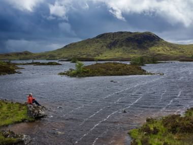 Wanderer in oranger Jacke sitzt auf Felsen am Wasser im Rannoch Moor, West Highland Way. Grüne Hügel und dramatischer Himmel im Hintergrund.