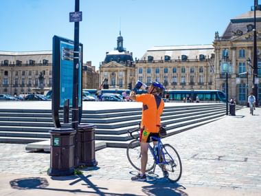 Cycliste en maillot orange vif avec casque bleu debout avec vélo sur place pavée à Bordeaux, architecture française classique en arrière-plan.