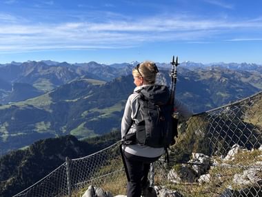Wanderin mit Rucksack und Wanderstöcken steht am felsigen Gipfel-Aussichtspunkt auf dem Stockhorn mit Blick auf Alpentäler und Bergketten.