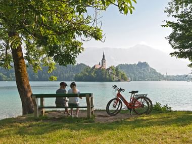 Radpause zweier Frauen auf einer Bank am Ufer des Bleder Sees mit Blick auf die Insel mit Kirche