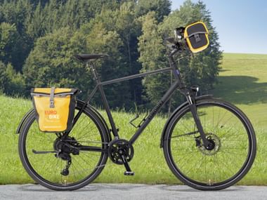 Schwarzes Tourenrad mit gelben Eurobike-Taschen und Lenkertasche, auf einer Straße vor grüner Wiese und Wald.