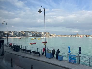 Uferpromenade in Cornwall mit Blick auf den Hafen von St Ives. Bunte Boote im türkisfarbenen Wasser, Küstenstadt am gegenüberliegenden Ufer.