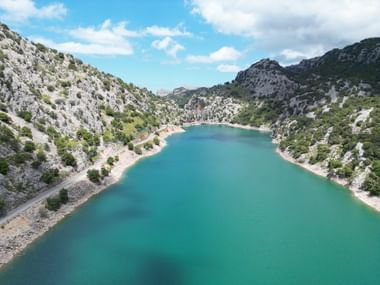 Türkisblauer Stausee Gorg, umgeben von Bergen mit Bäumen. Links davon verläuft eine Strasse.
