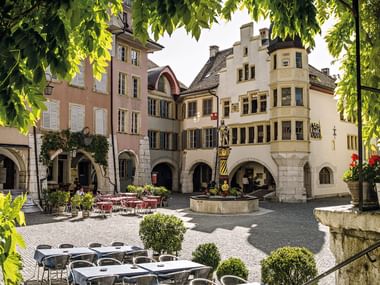 Charmante place pavée à Bienne entourée de bâtiments historiques avec arcades, terrasses de café et une fontaine centrale sous les arbres.