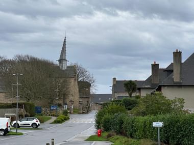 Dorfstraße in Plougrescant, Bretagne, mit dem markanten gedrehten Turm der Kapelle Saint-Gonéry hinter Steinmauern und Häusern.