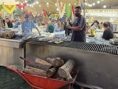 Großer Außengrill auf dem Feiras Novas Festival in Ponte de Lima. Ein Koch betreut den Grill, während Besucher unter bunten Dekorationen speisen.