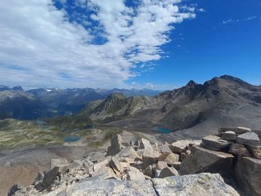 Felsiger Gipfel mit Steinmännchen mit Blick auf alpine Landschaft mit Bergseen, Gipfeln und Tälern unter blauem Himmel mit weißen Wolken.