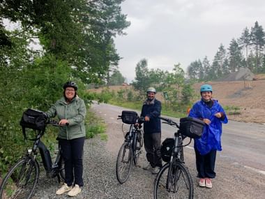 Drei Radfahrer mit beladenen Tourenrädern auf einer Landstraße bei Mariefred. Wald und bewölkter Himmel im Hintergrund.