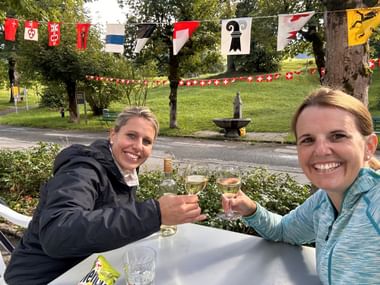 Zwei lächelnde Frauen stoßen mit Weißwein an einem Tisch im Freien an. Schweizer Kantonsflaggen und Schweizer Kreuz-Wimpel schmücken den Hintergrund mit Brunnen.