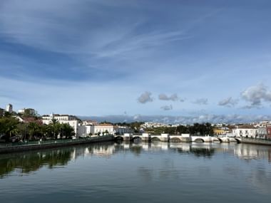 Weiße Ponte Romana Brücke mit mehreren Bögen über ruhigem Fluss in Tavira, Algarve. Historische Gebäude und Palmen säumen das Ufer unter blauem Himmel.