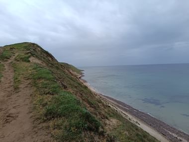 Wanderweg entlang grasbewachsener Klippen mit Blick auf die Ostsee zwischen Ystad und Kåseberga am Österlenleden. Grauer bewölkter Himmel über ruhigem Wasser.