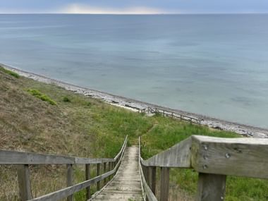 Holztreppe mit Geländer führt einen grasbewachsenen Hang hinunter zu einem Kieselstrand. Die Ostsee erstreckt sich bis zum Horizont unter bewölktem Himmel.