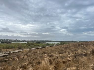 Holzsteg durch Sanddünen zur Lagoa dos Salgados Lagune mit grüner Vegetation und bewölktem Himmel in der Algarve.