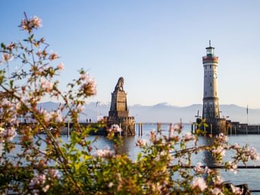 Lindauer Hafeneinfahrt am Bodensee mit weißem Leuchtturm und Bayerischem Löwen, gerahmt von blühenden Ästen im Vordergrund.