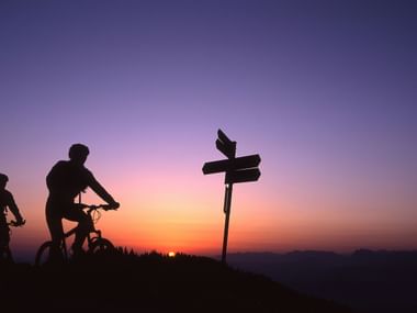 Zwei Mountainbiker radeln bei Sonnenuntergang an einem Wegweiser vorbei.