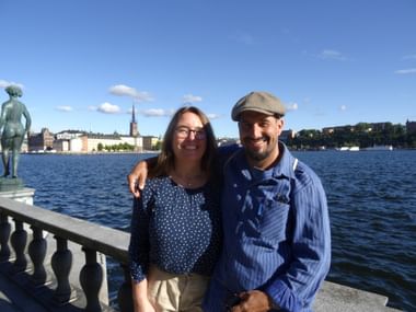 Lächelndes Paar an einer Balustrade am Wasser mit Stockholms Altstadtskyline und Kirchturm im Hintergrund unter blauem Himmel.