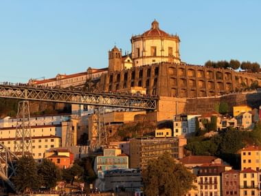 Sonnenuntergang in Porto mit historischem Kloster auf dem Hügel, Dom Luís I Brücke und bunten Gebäuden in goldenem Licht am Flussufer.