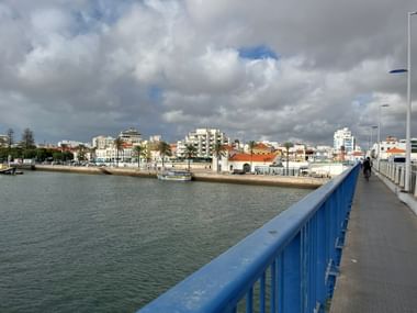 Fußgängerbrücke mit blauem Geländer über den Arade-Fluss in Portimão, Algarve. Blick auf Uferpromenade mit Palmen und weißen Gebäuden.