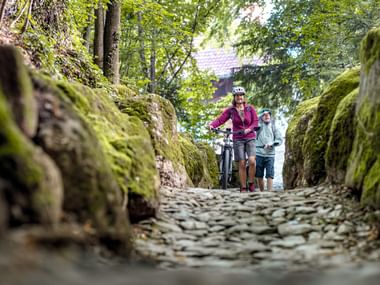 Zwei Mountainbiker schieben ihre Räder durch die schmale Strasse im Wald.