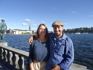 Lächelndes Paar an der Uferpromenade mit Stockholms Altstadt-Skyline und Kirchturm über blauem Wasser unter klarem Himmel.