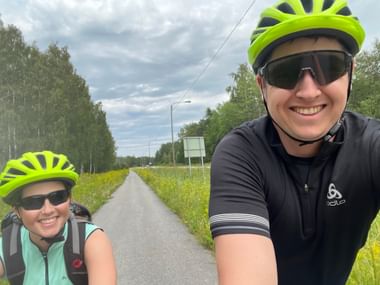 Zwei Personen sind auf einem Fahrradweg. Beide tragen ein Fahrradshirt, einen gelb-grünen Helm und eine Sonnenbrille.