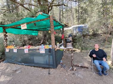 Mitten im Wald gibt es einen grünen Stand der an einen Kiosk erinnert. Ein Mann daneben in einem Stuhl