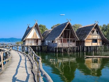 Historische Pfahlbauten in Unteruhldingen am Bodensee mit Holzsteg zu rekonstruierten Stelzenhäusern über klarem Wasser.
