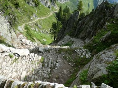 Die berühmten "Steigle" vor der Massaschlucht. Schmale Steinstufen führen steil durch felsiges Gelände am Aletsch-Panoramaweg hinab, mit grünen Almwiesen und Bergen im Hintergrund.