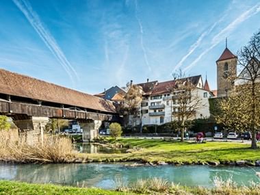 Das Schmuckstück Aarberg im Berner Seeland. Überdachte Holzbrücke über die Aare in Aarberg, mit historischen Gebäuden und einem Kirchturm im Hintergrund unter blauem Himmel.