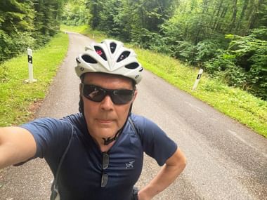 Radfahrer Werner macht Selfie auf Waldstraße mit weißem Helm und Sonnenbrille. Asphaltstraße führt durch üppigen grünen Wald.