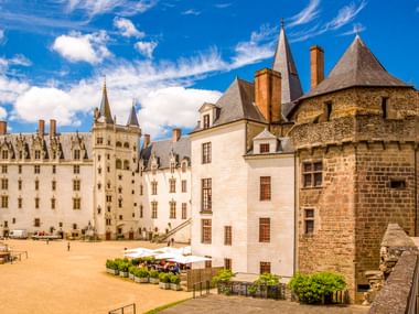 Château des Ducs de Bretagne à Nantes avec murs en pierre blanche, tour ronde et cour avec étals de marché sous ciel bleu.