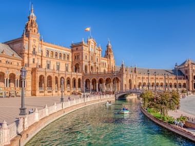 Plaza de España in Seville with ornate brick buildings, arched colonnade, and canal with bridge under clear blue sky.