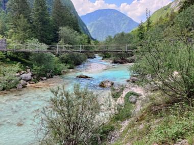 Eine Hängebrücke über dem türkisblauen Fluss Soca, Slowenien. Umgeben von bewaldeten Bergen und Bäumen,