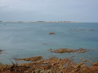 Ruhiges blaues Meer mit Felsformationen bei Tréguier, Bretagne. Entfernte Inseln und ein Leuchtturm am Horizont unter bewölktem Himmel.