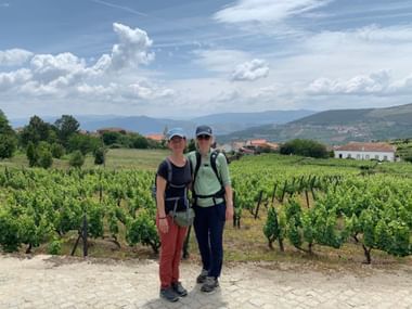 Zwei Wanderinnen mit Rucksäcken in Weinberg bei Vilarinho de São Romão. Grüne Reben, Hügel und weiße Gebäude unter blauem Himmel.