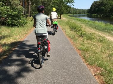 Zwei Radfahrer mit Packtaschen fahren auf asphaltiertem Radweg durch mecklenburgische Landschaft. Fluss fliesst neben Weg mit dichtem Wald.