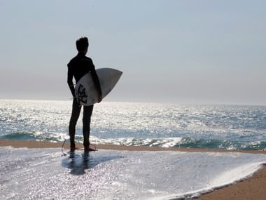 Silhouette eines Surfers im Neoprenanzug mit weißem Surfbrett, stehend auf nassem Sand am Strand von Ericeira mit glitzernden Wellen.