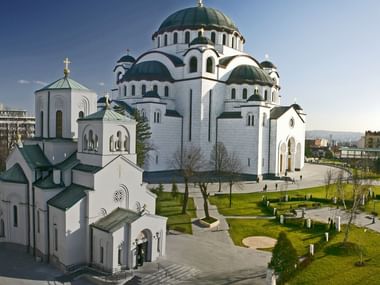 Große weiße orthodoxe Kathedrale mit grünen Kuppeln und goldenen Kreuzen in Belgrad, umgeben von Grünanlagen und städtischen Gebäuden.
