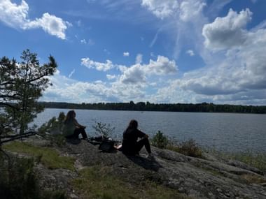 Zwei Personen sitzen auf felsigem Ufer mit Blick auf einen ruhigen See mit Wald in der Ferne unter blauem Himmel mit weißen Wolken.