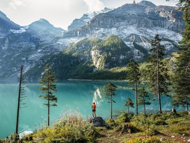 Randonneur en veste rouge debout sur un promontoire rocheux surplombant le lac turquoise d'Oeschinen entouré de sommets alpins enneigés.