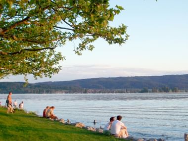Menschen entspannen am grasbewachsenen Seeufer in Radolfzell bei Sonnenuntergang. Der Bodensee erstreckt sich zu bewaldeten Hügeln unter weichem Abendhimmel.