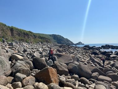 Wanderer überqueren einen felsigen Strand in Cornwall mit großen runden Felsbrocken. Ein Regenbogen erscheint am blauen Himmel über den Küstenklippen.