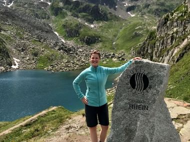 Alexandra am Rheinquelle-Steinmarker Alexandra in türkisfarbener Jacke posiert neben Rheinquelle-Steinmarker mit Bergsee und Alpengipfeln im Hintergrund.