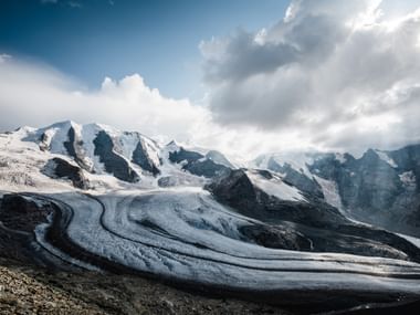 Ein atemberaubendes Bild des Diavolezza Gletschers der in der Mitte in einer Kurve vom Schneebedeckten Berg herab führt. Rechts oben sind Wolken in verschiedenen grautönen die links in der oberen Ecke noch etwas blauen Himmel hervor schauen lassen.