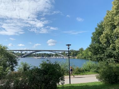 Bogenbrücke über Wasser mit Booten, gesehen von grünem Uferweg. Bäume rahmen die Szene unter blauem Himmel mit weißen Wolken.