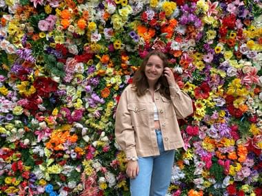 Riana Achermann vor bunter Blumenwand Riana Achermann steht vor einer lebendigen Blumenwand, trägt eine beige Jacke und blaue Jeans, lächelt und berührt ihr Haar.