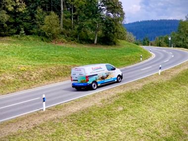 Weißer Transporter mit bunter Gepäcktransport-Beschriftung fährt auf kurvenreicher Landstraße durch grüne Hügel und Wälder unter bewölktem Himmel.